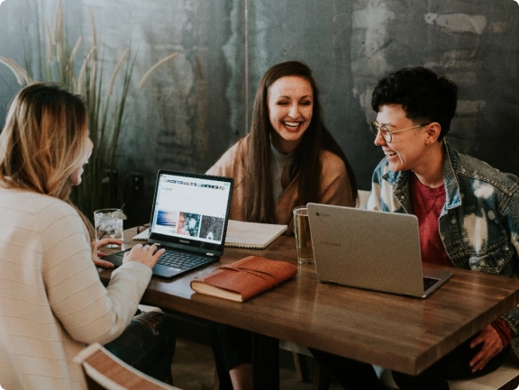 A photograph of a customer interaction where 3 people are sat around a table, working on laptops and laughing